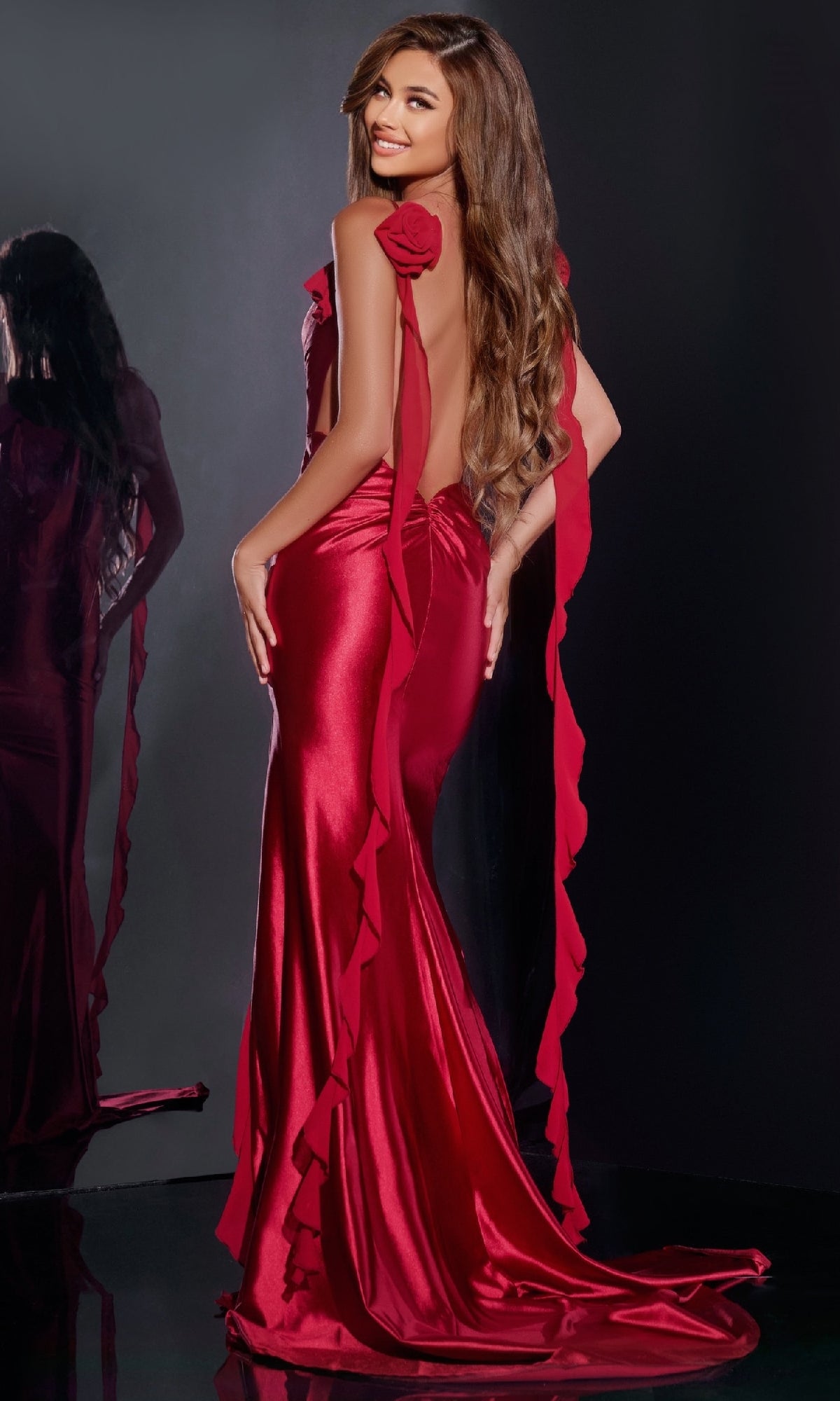 A woman in the Jovani D5078 long prom dress—a shiny red backless satin gown with a 3D flower neckline—stands smiling with long wavy hair, her reflection visible in a mirror against a dark background.